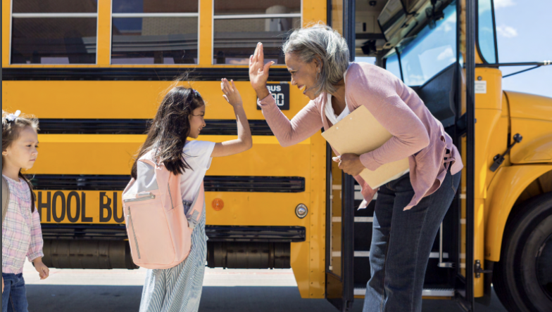 A child and adult in front of a school bus, giving each other a high five.