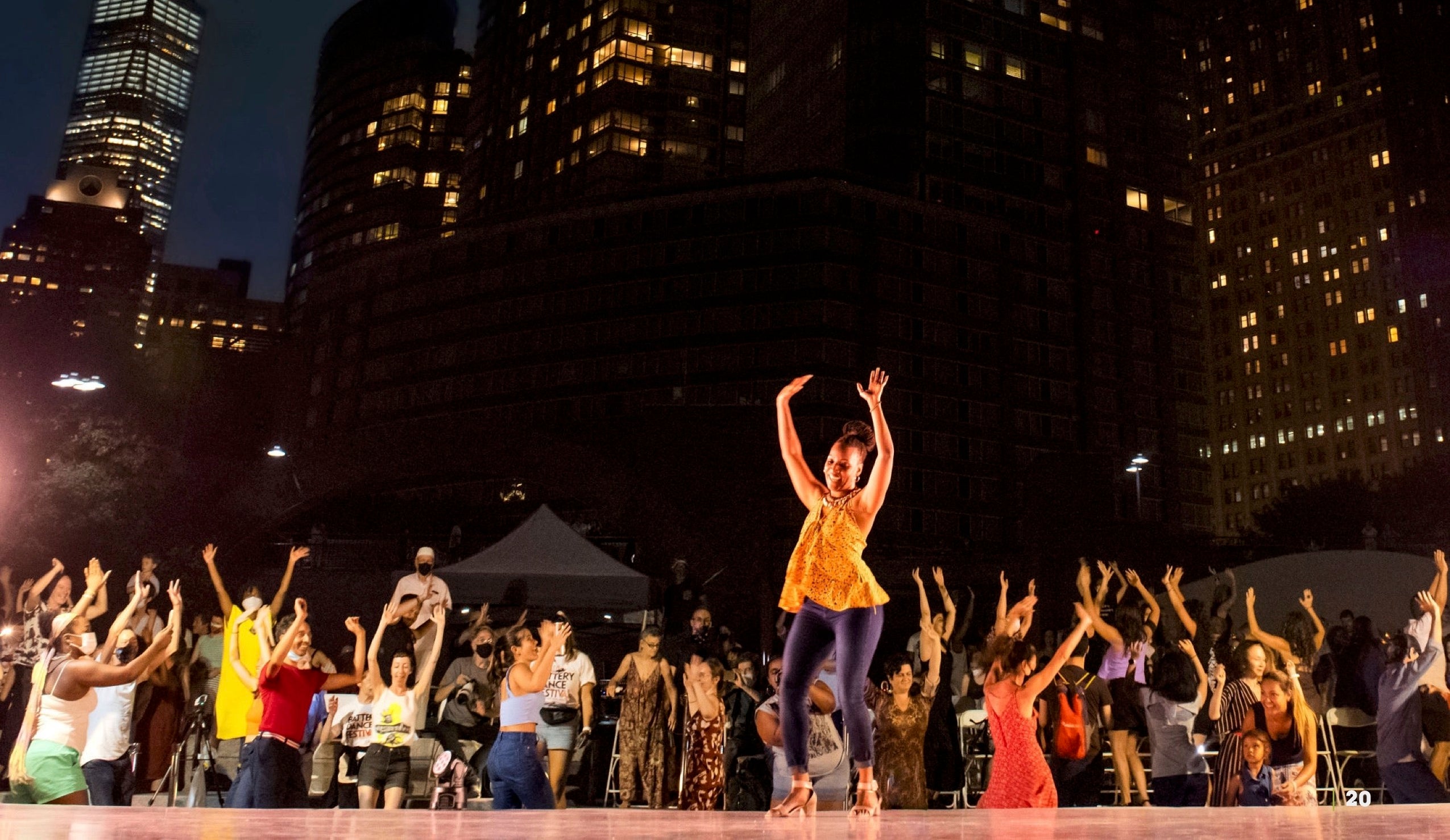 A Black woman dances on stage in front of high-rise buildings while her community cheers her on.