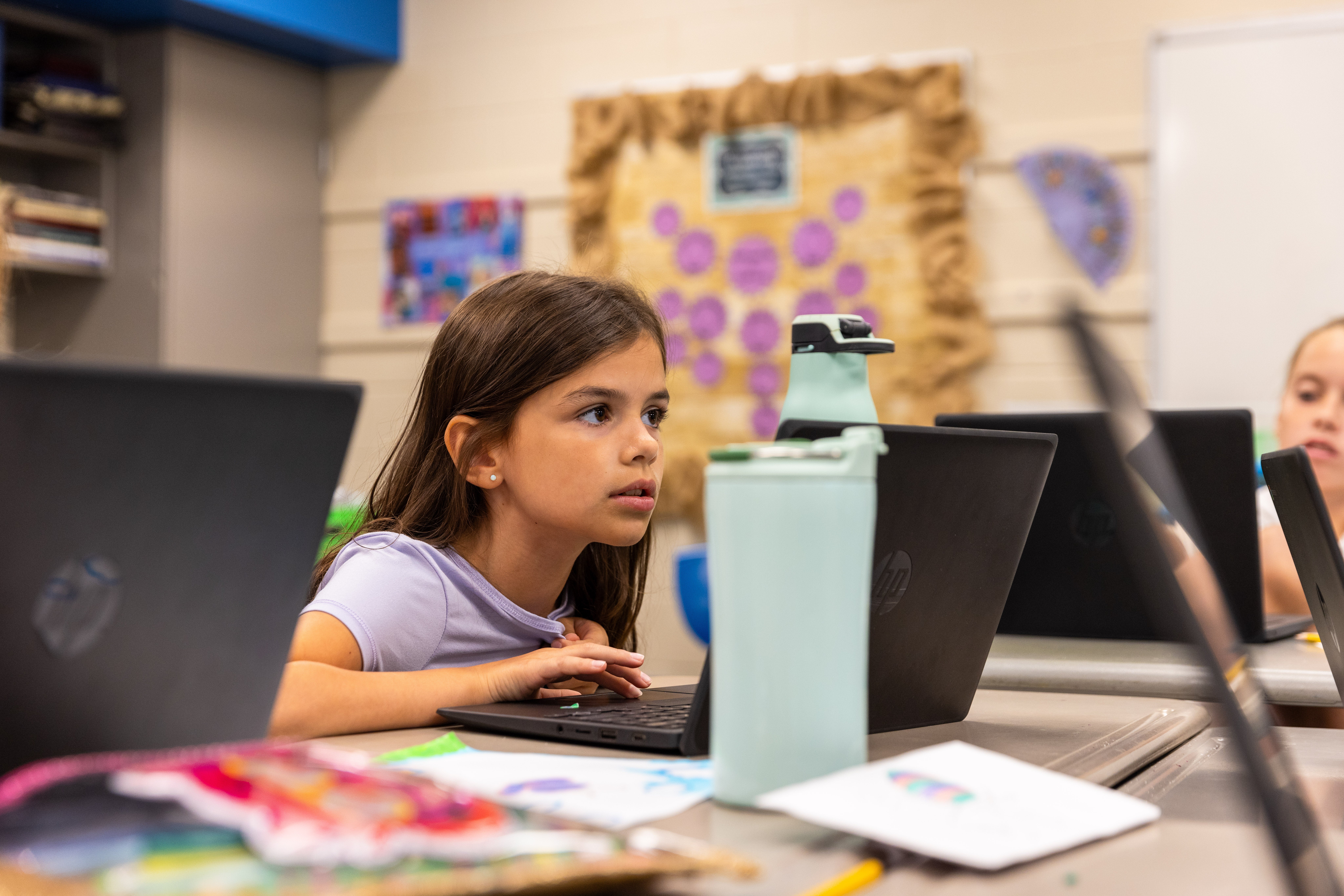 A young girl sits at a desk looking at the screen on her laptop.