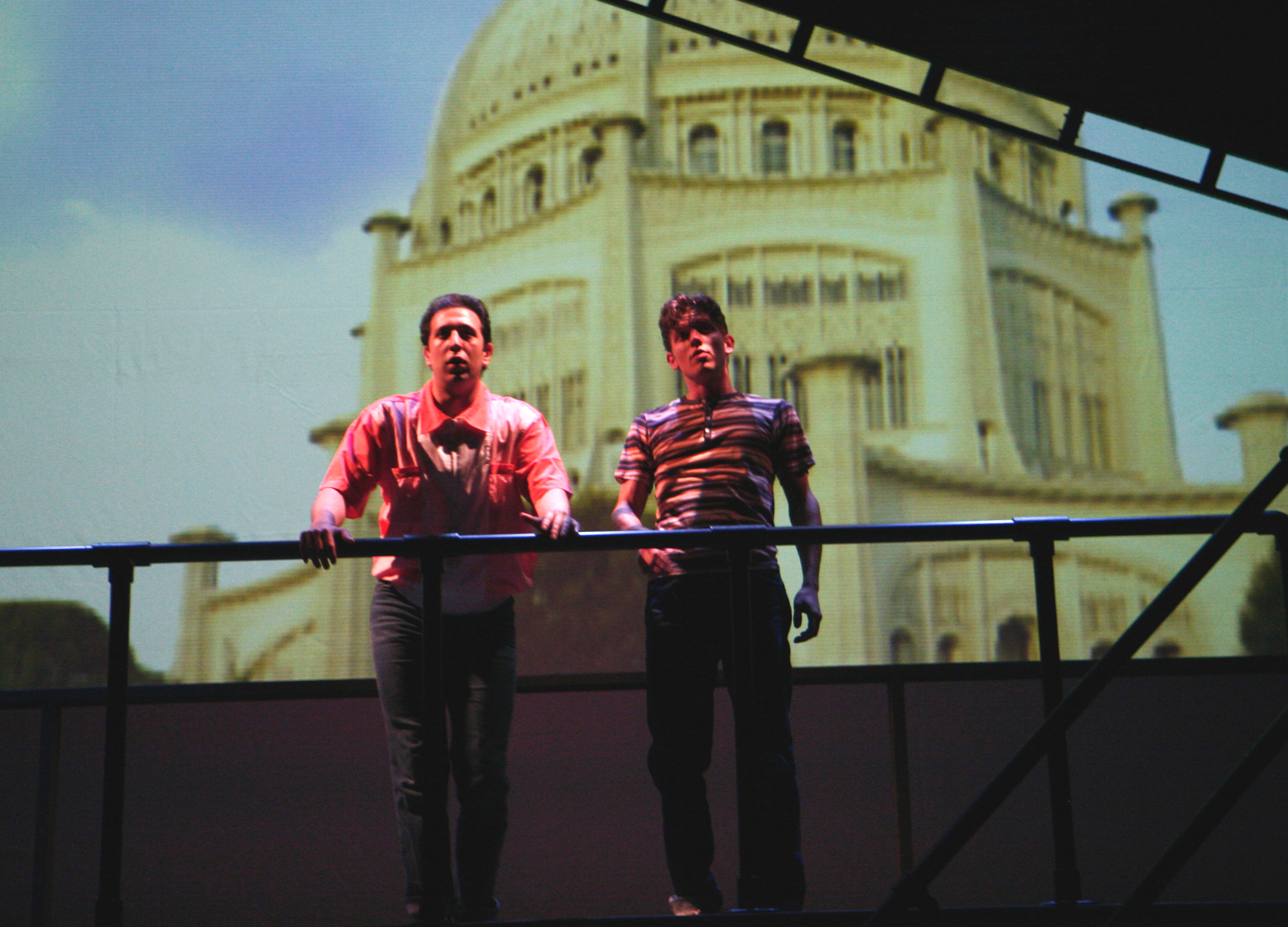 Two actors stand by a railing against a backdrop of a grand building.