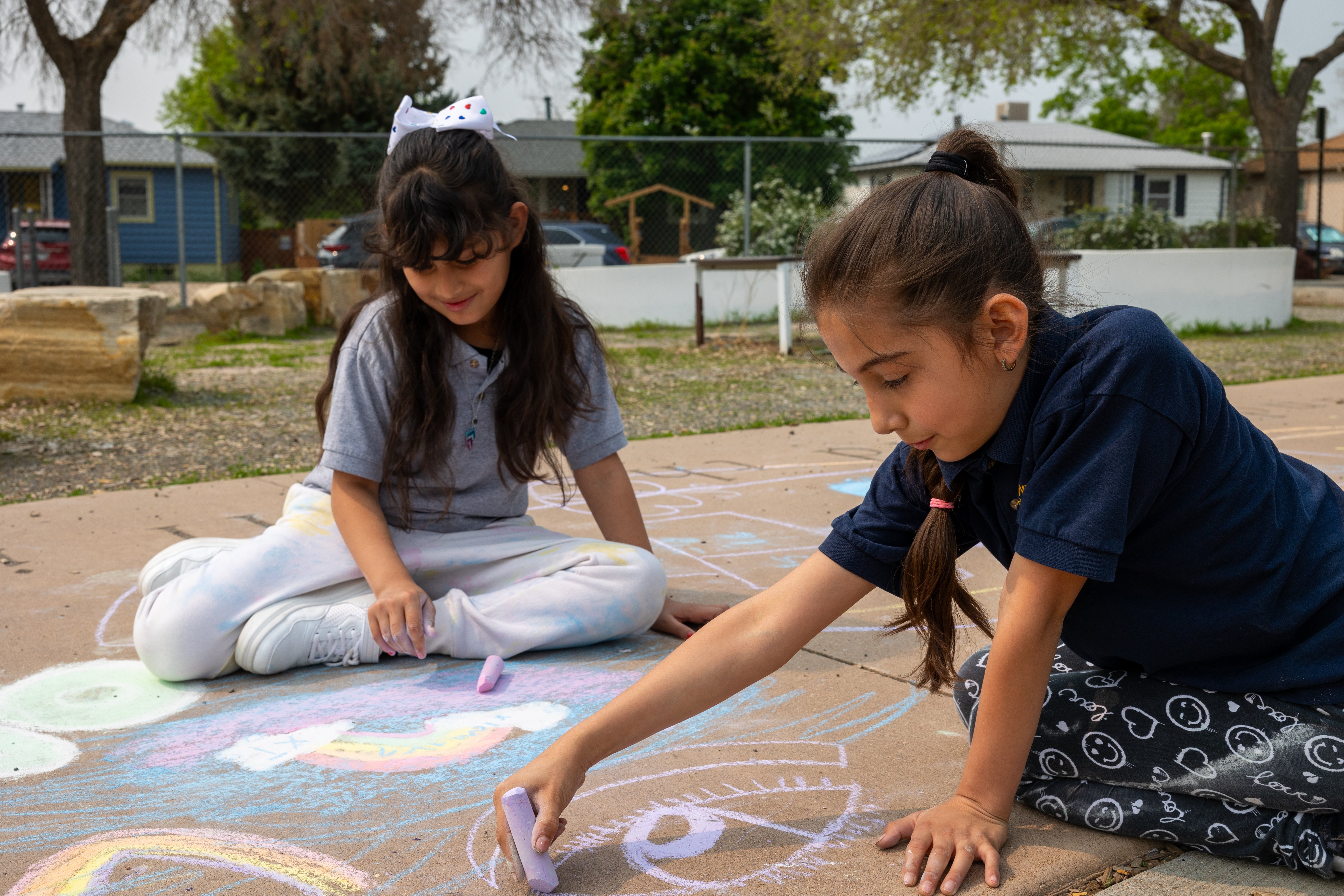 Two girls draw with sidewalk chalk outside.