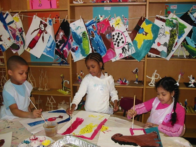 Three children work on their paintings in front of a string of drying art.