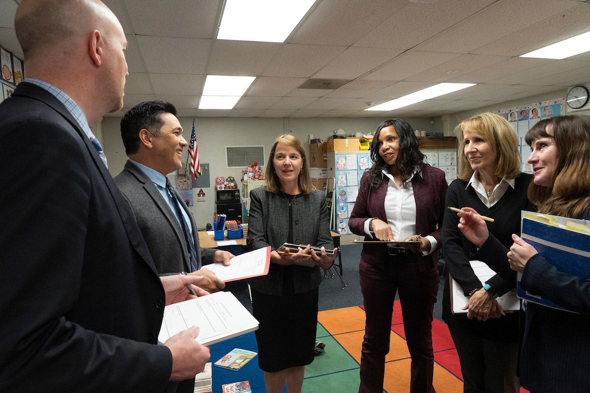 A group of education leaders with notepads stand in a discussion circle in a classroom.