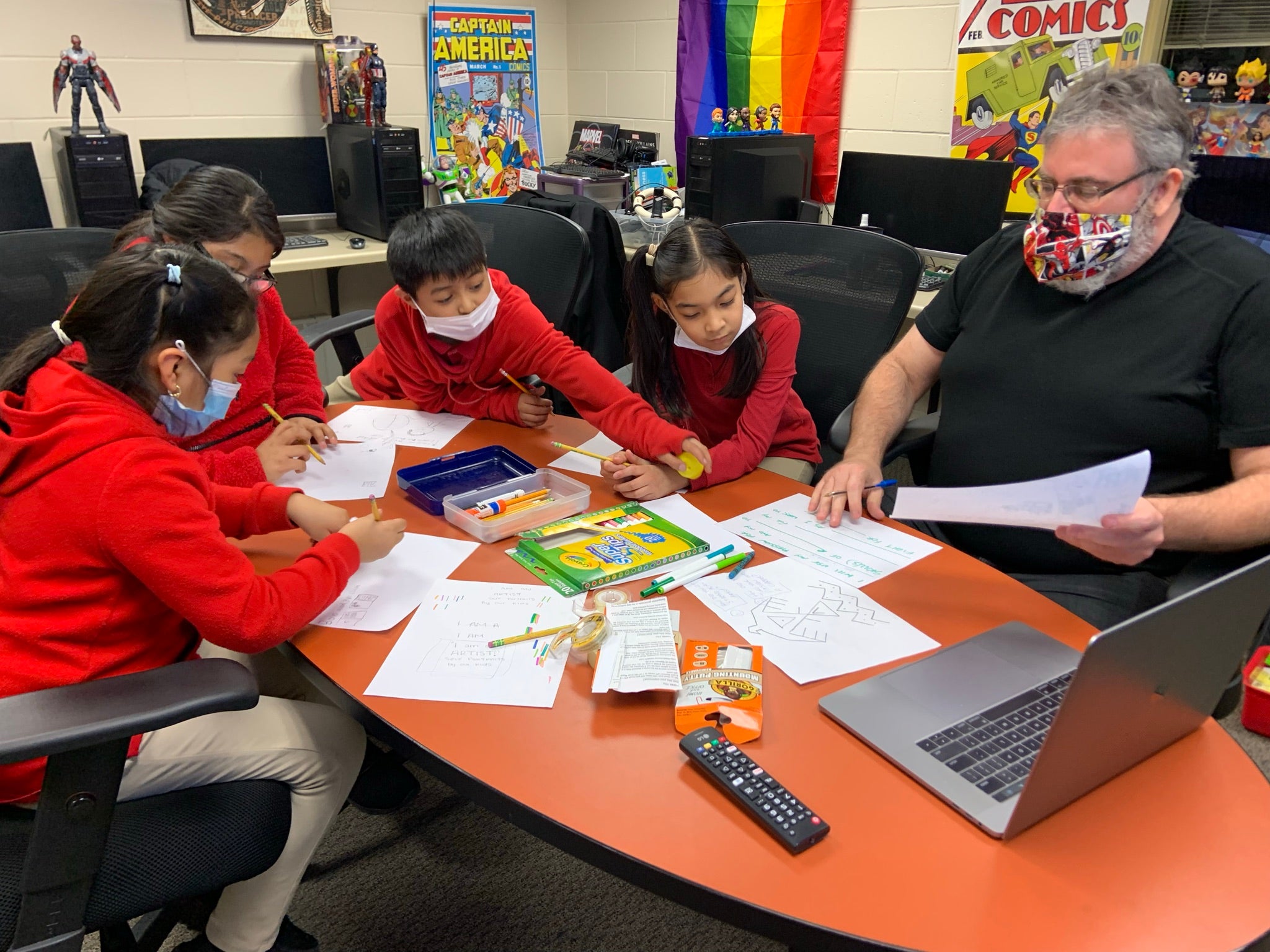 Students wearing masks sit around a table scattered with worksheets.