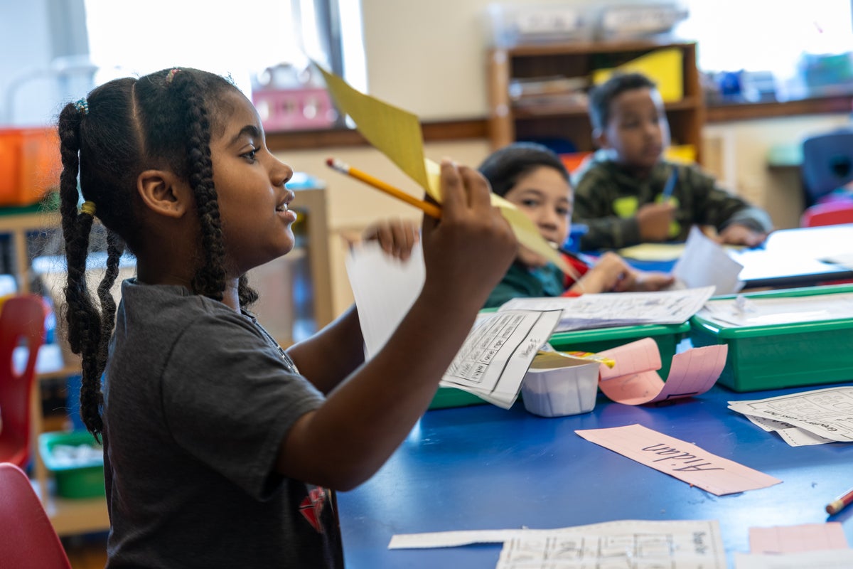 Young students in classroom setting, two are black boys and one black girl with long hair in foreground holding paper and pencil