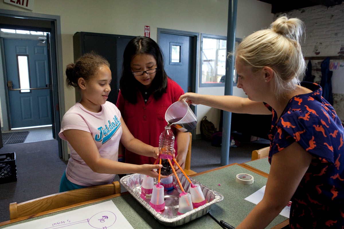 Female white blond teacher helps two girls with brown skin and hair with science experiment