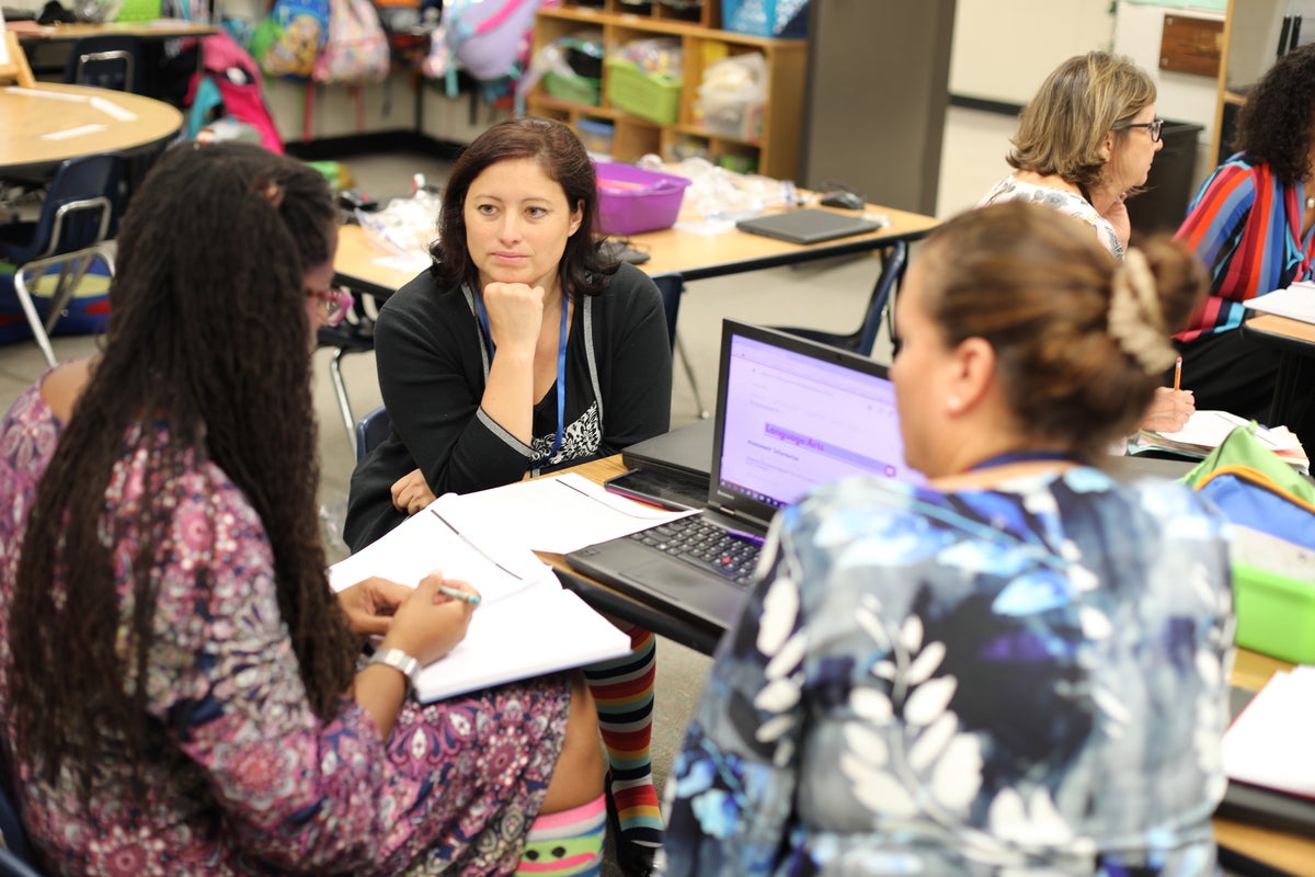 3 adult women sitting around desks with laptops talking to each other