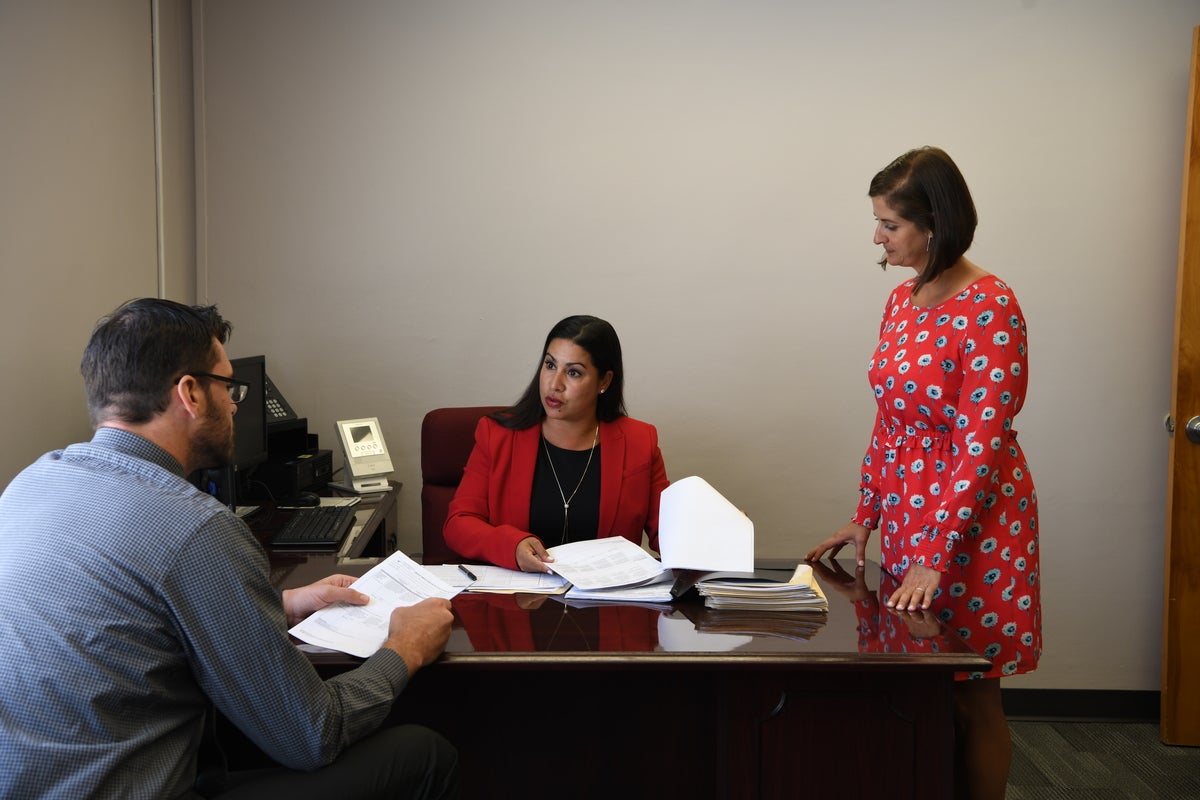 2 female adults and 1 male adult sit around desk looking at each other. books and papers on desk. one woman is standing up.