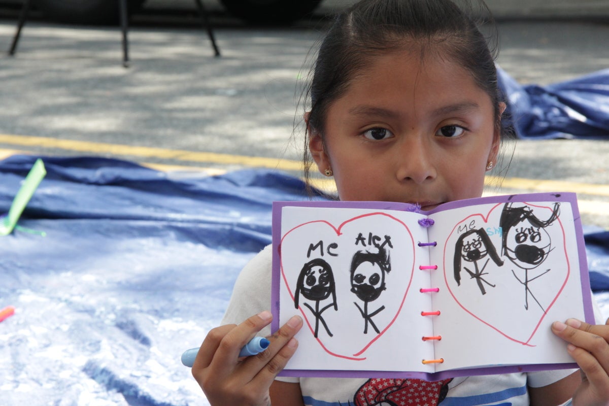 Hispanic elementary school aged girl holding up book with drawings of stick figures in hearts