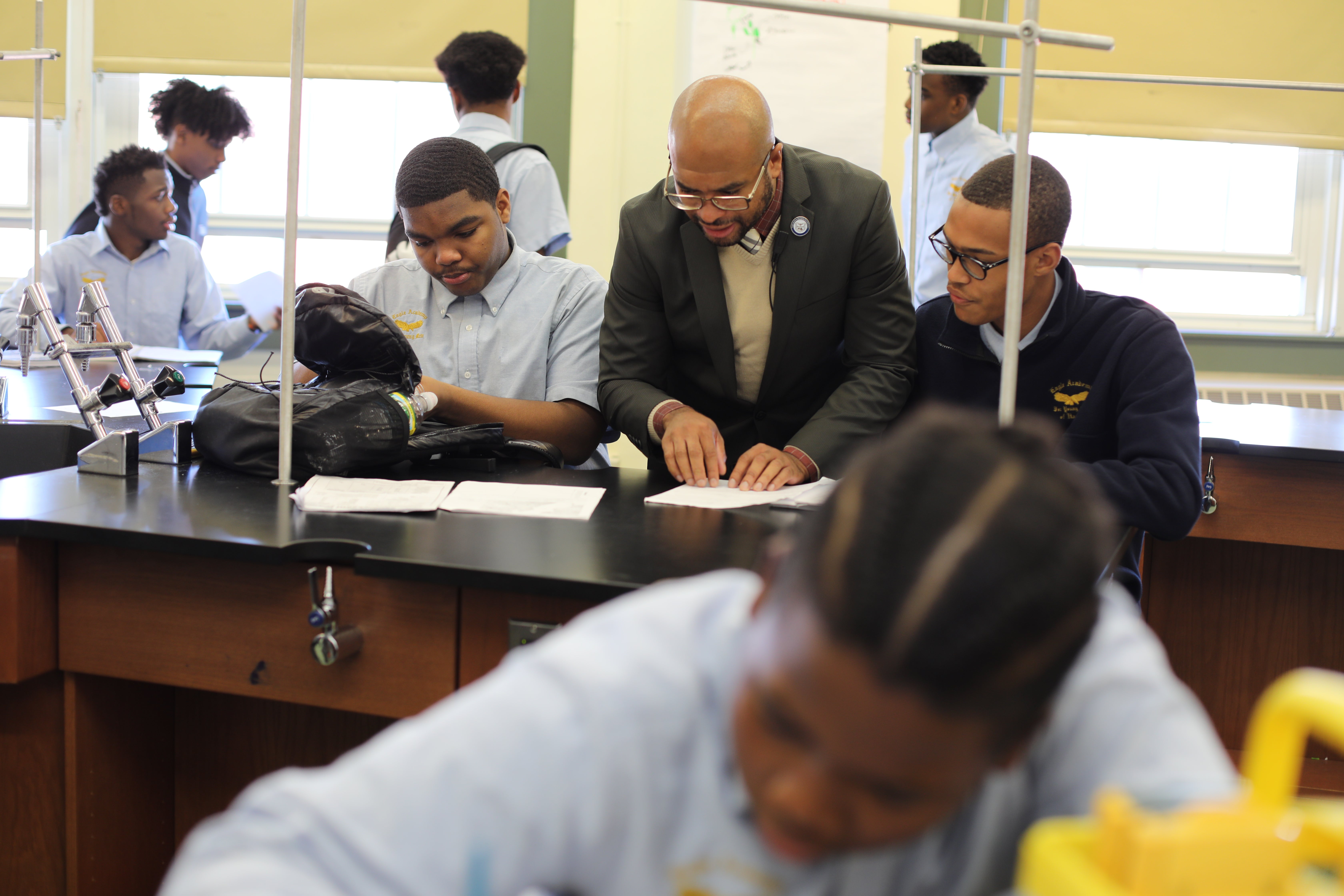 A Black principal wearing a suit is assisting students in a classroom. The students are seated at a lab table, engaged in reading or writing activities. The classroom has windows and science-related equipment visible.