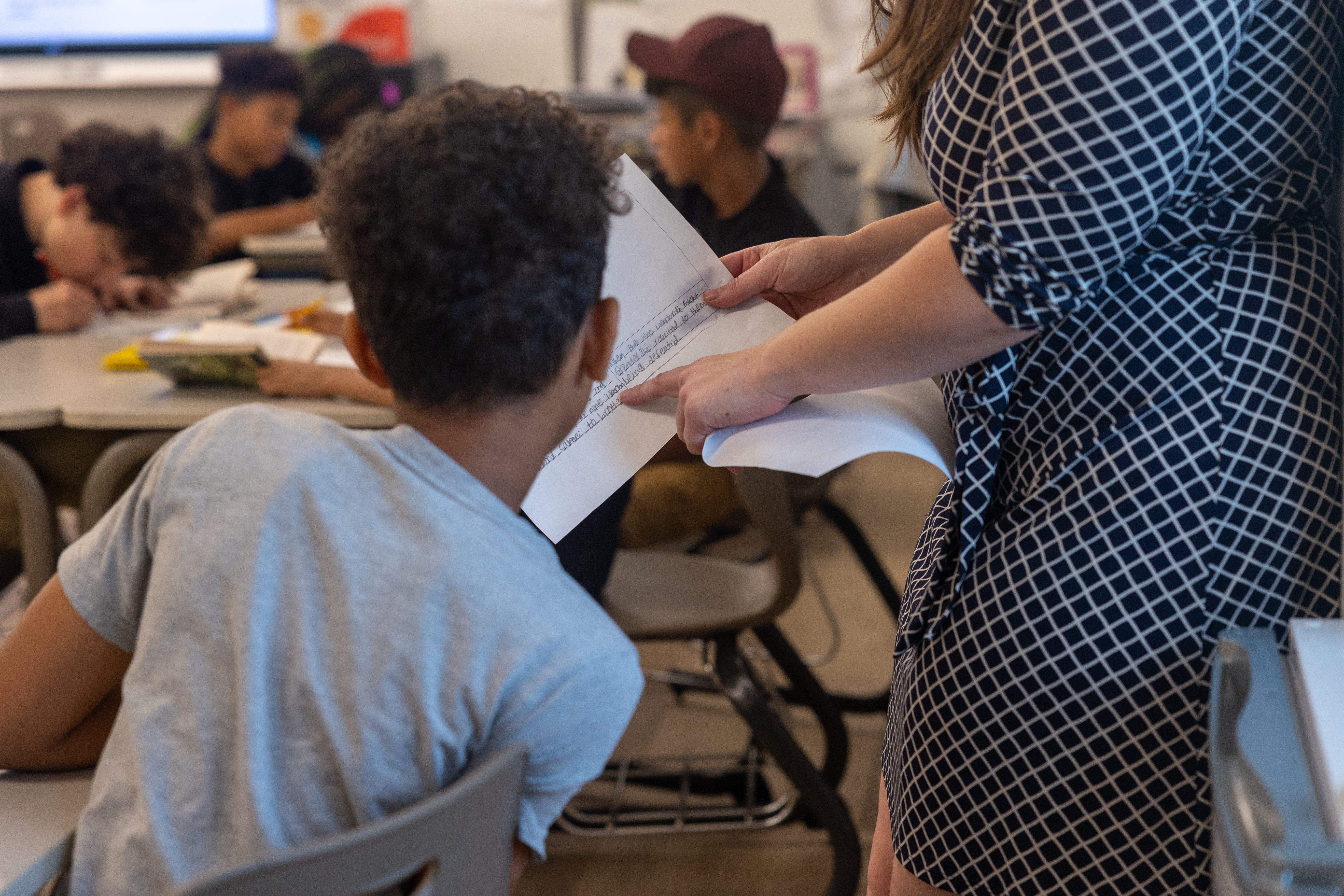 An educator stands next to a student who is sitting down at a desk in a classroom. They are both looking at a piece of paper. There are students in the background working.