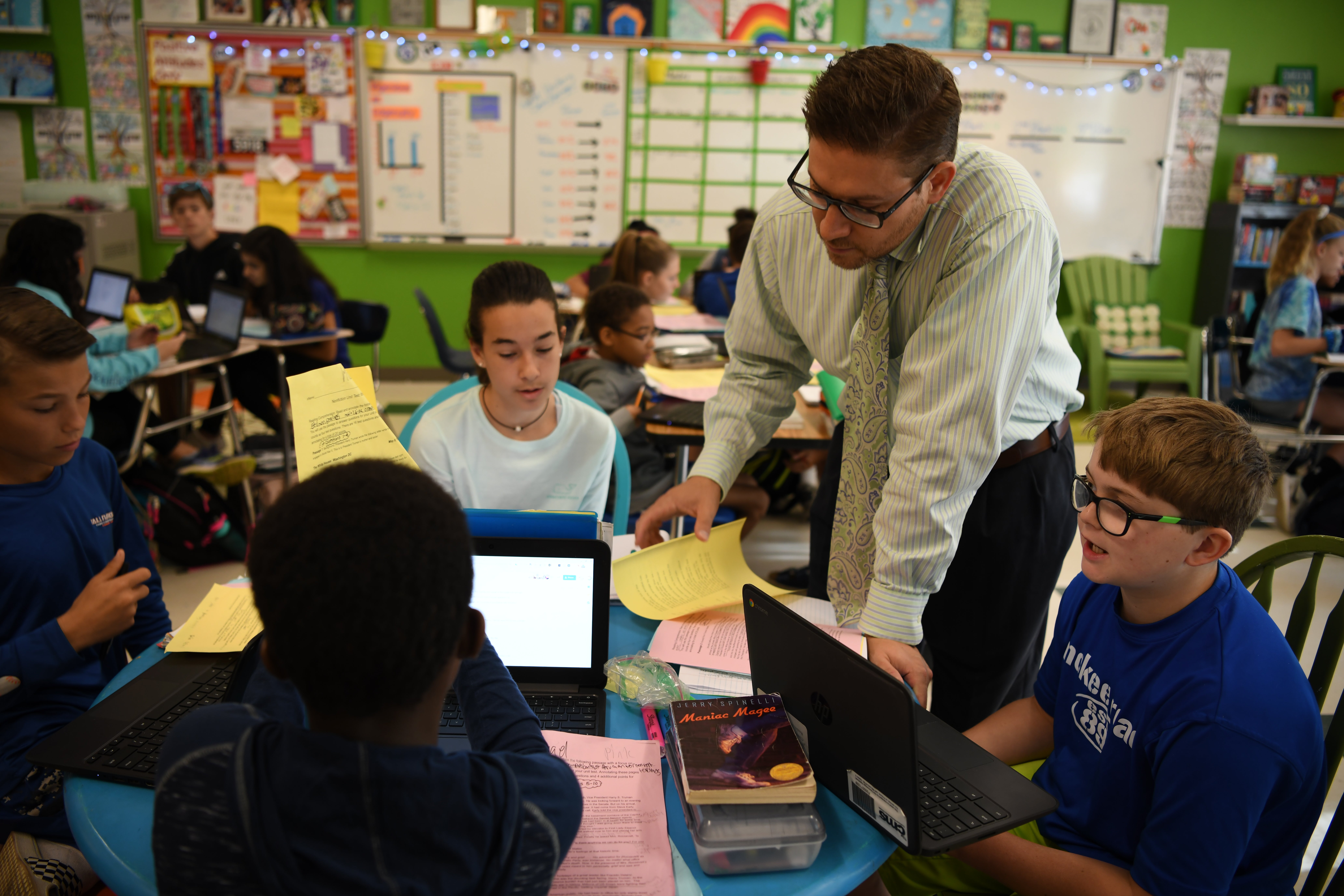 A group of students working on laptops in a classroom, with an educator leaning over to assist them. The classroom has colorful charts and a lively atmosphere.