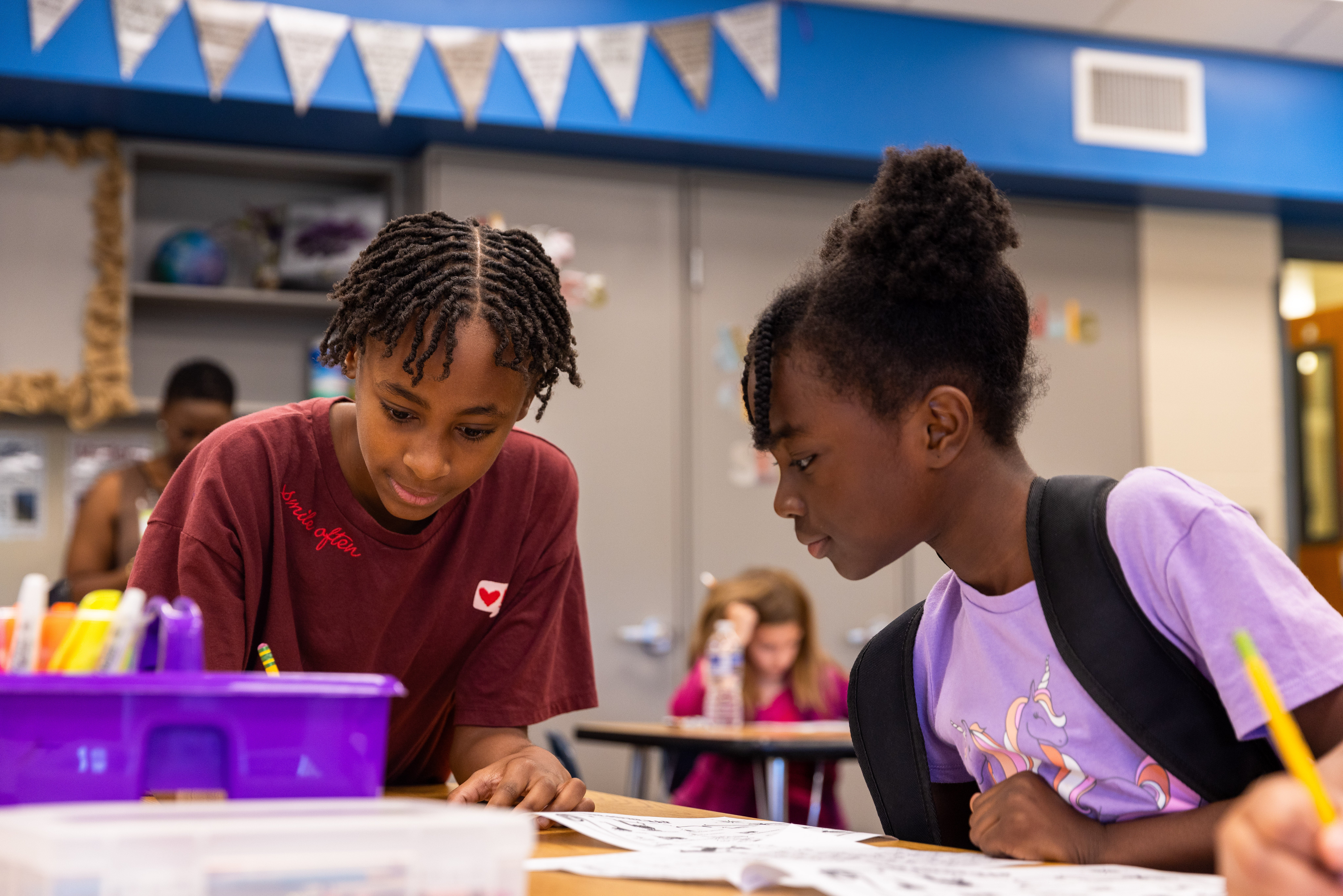 Two students are engaged in classroom work, focusing on papers at a table. One is wearing a maroon t-shirt and the other a lavender t-shirt and a black backpack. The classroom has bright walls and a variety of supplies, including a purple organizer filled with writing tools. In the background, other students are also working.