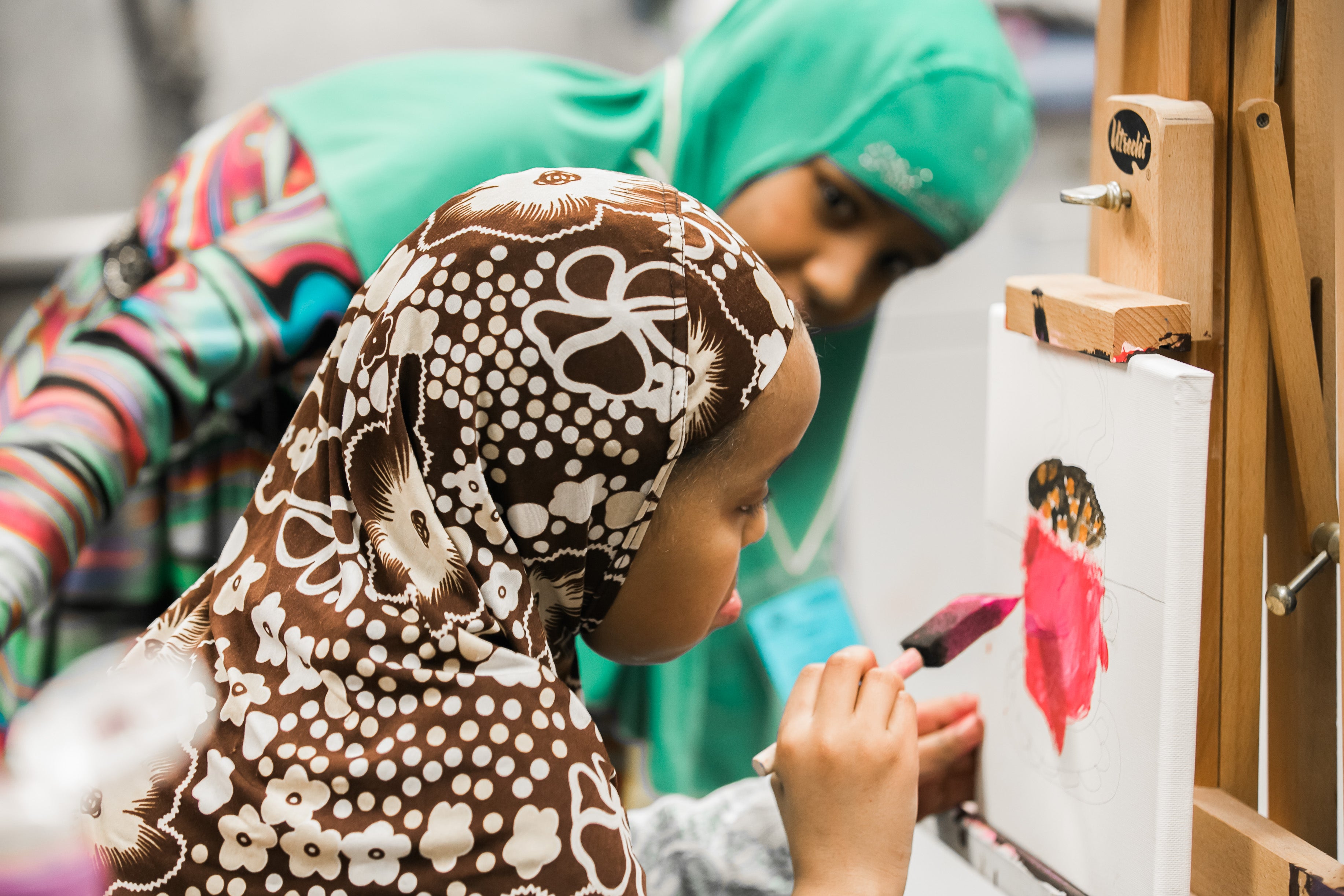 Two people are painting at an easel. One is painting on the canvas, while the other observes closely. They are wearing brightly patterned clothing and headscarves.