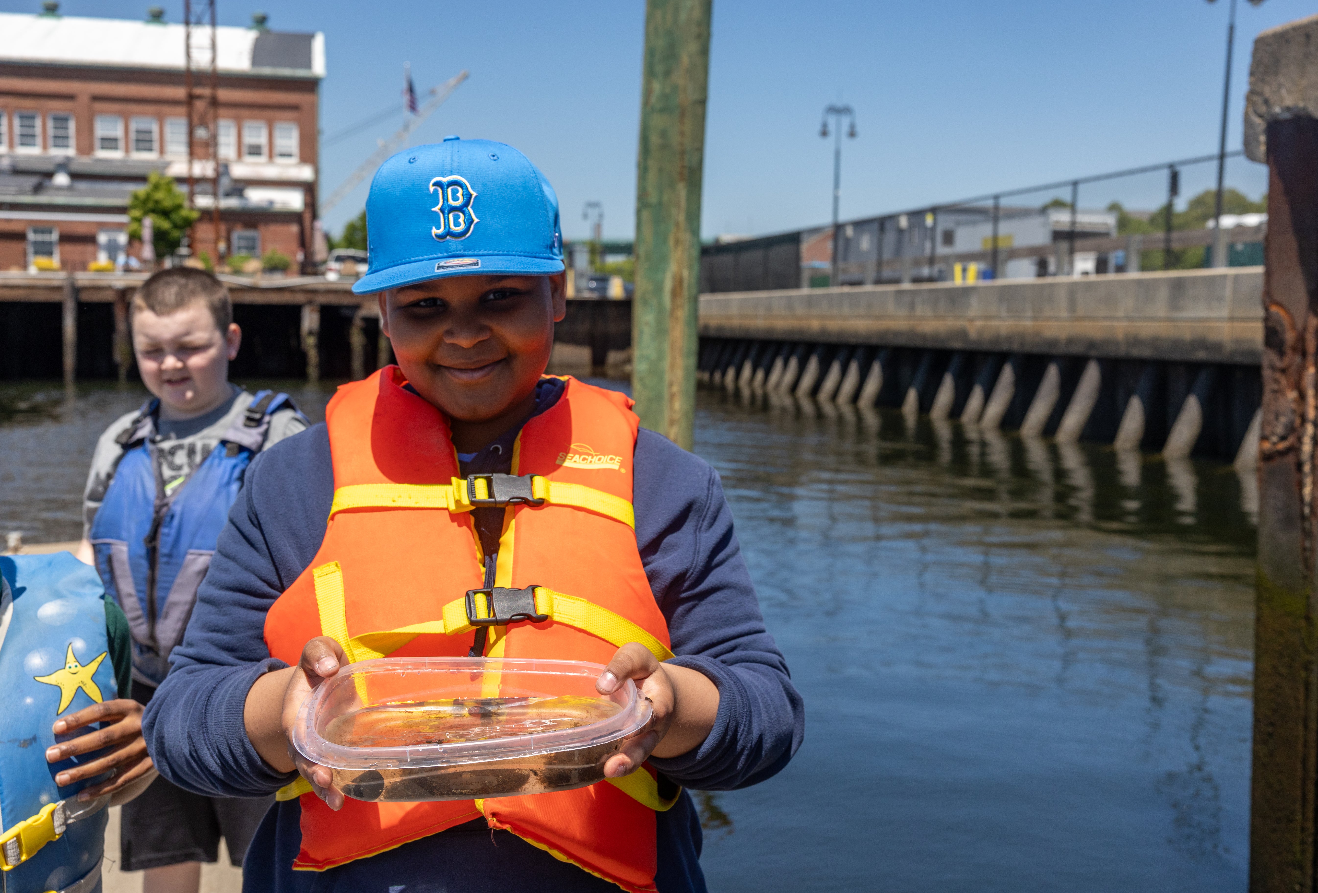 Young boy is standing on a dock holding a container filled with water. He is wearing an orange life jacket and blue hat. There is another boy standing behind him.