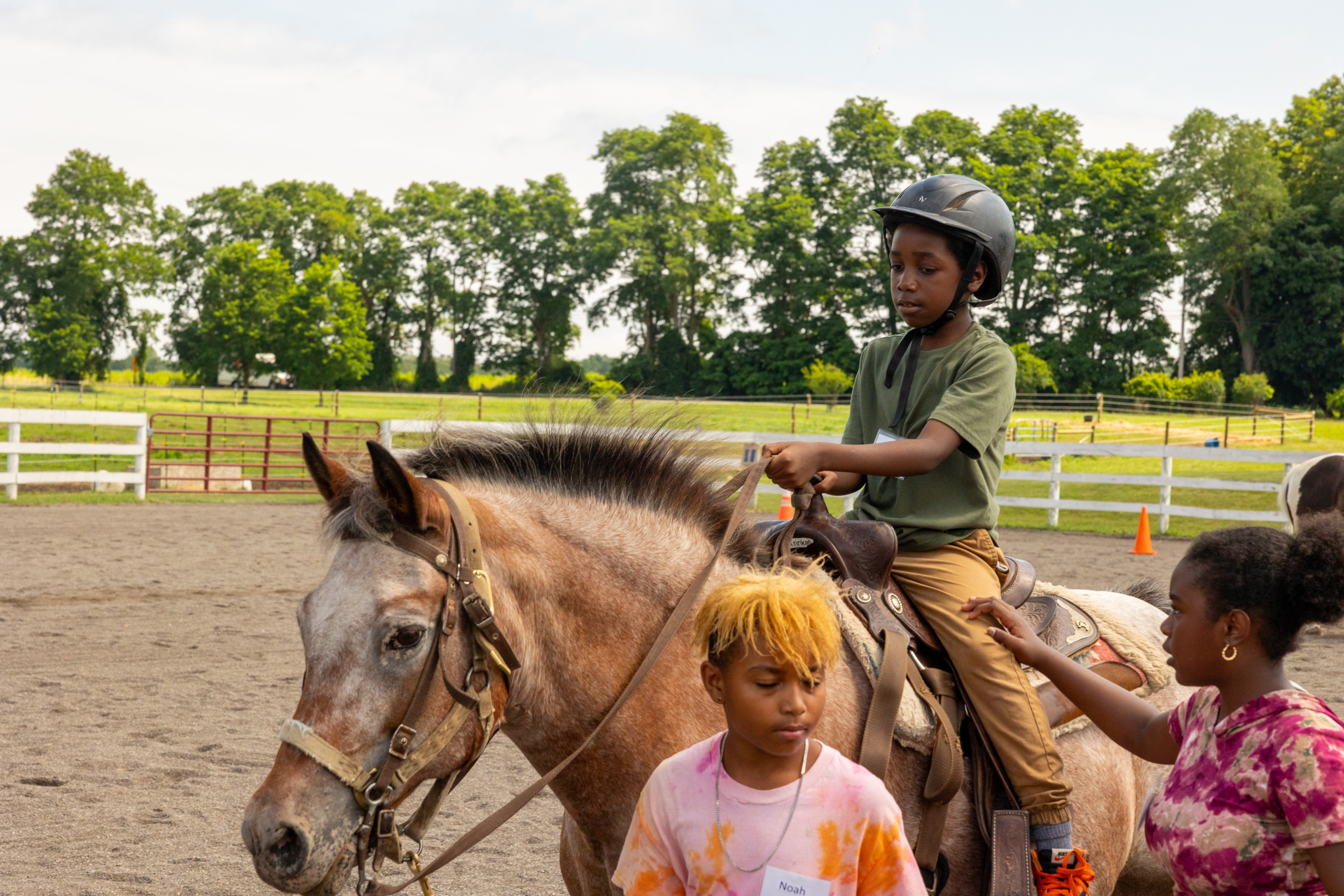 A young boy wearing a green shirt and helmet rides a brown horse in a field while an instructor helps support him and another student stands in front of the horse