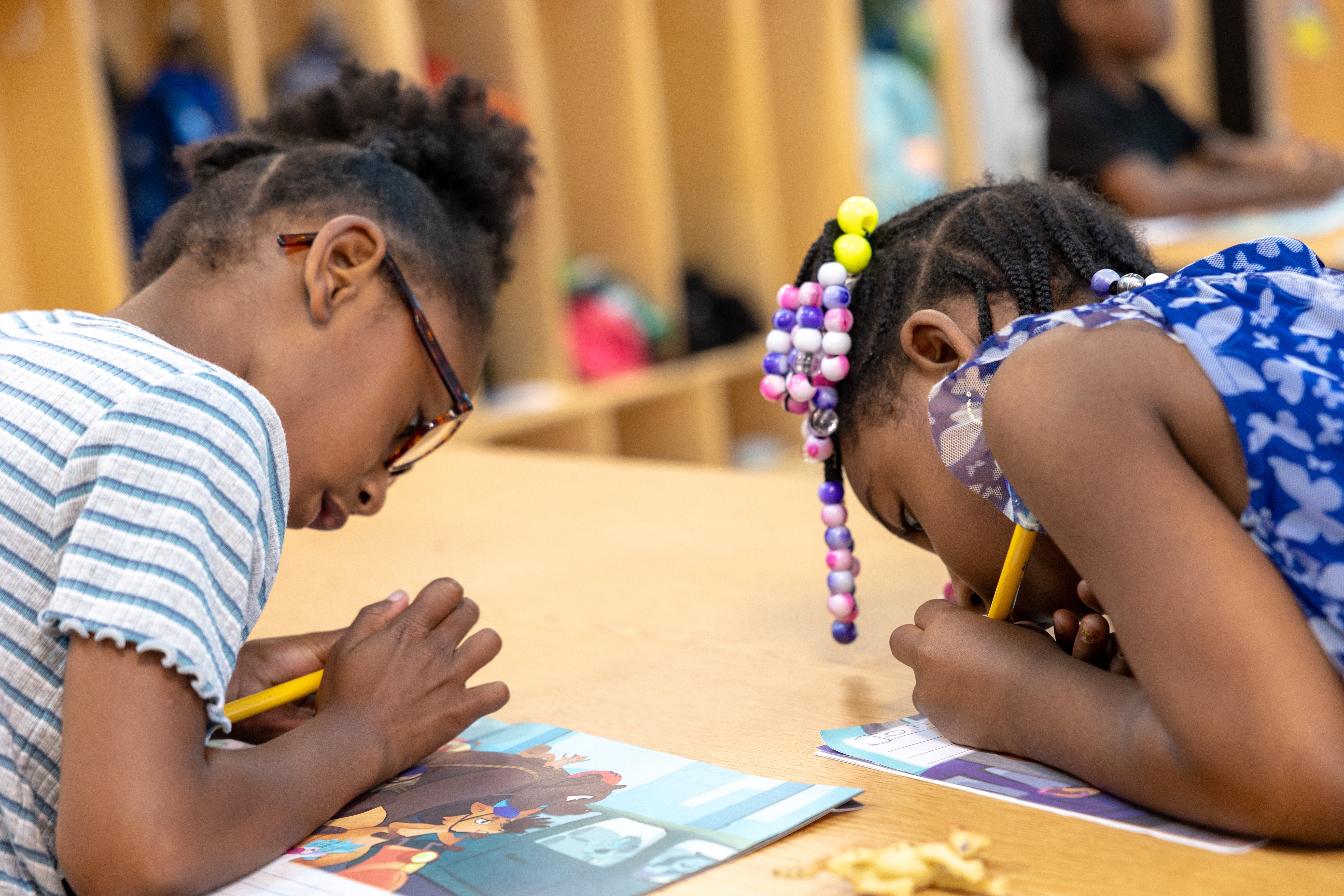 Two young girls are drawing with pencils while sitting at a desk in a classroom