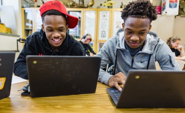Two teenage boys sit at a classroom table using laptops at their summer learning program, smiling and focused on their screens.