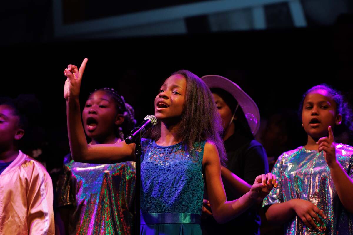 A group of young black girls singing on stage in a middle school