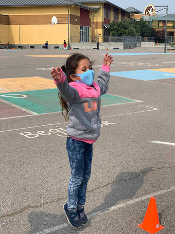 Little girl wearing a face mask jumping in a school yard with her hands up. 